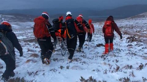 Rescuers wearing winter walking gear and helmets carry a stretcher over a snowy mountain landscape.