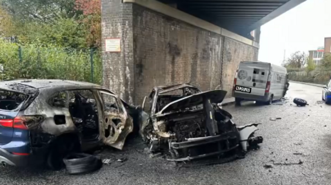 Two burnt out cars sit underneath a railway bridge. Both have suffered extensive damage and look completely destroyed. The van is parked a few feet away and has suffered fire damage, although it is less severe.