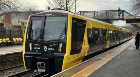 A Merseyrail black and yellow electric train on the track. A bridge over the platform can be seen above the train with a man walking across the other side. One man can be seen standing on the train platform at the bottom looking towards the train.