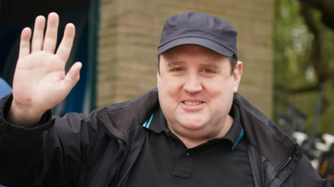 Kay is pictured waving at the camera and wearing a dark cap and top and jacket. He is outside in a street.