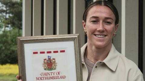 Lucy Bronze smiling and holding her official honour, which is a framed large document