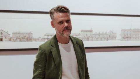 A stylish middle-aged man with a short greying beard and gelled hair poses in front of a large drawing of a streetscape.