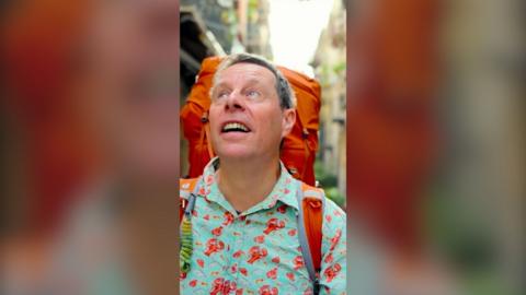 A man wearing a turquoise shirt with red lobsters on and an orange backpack looks up to the sky as he walks down a street.