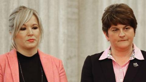 Michelle O'Neill standing next to Arlene Foster. O'Neill has blonde hair tied back with a fringe wearing a pink blazer and black top, and a long silver necklace. She is wearing makeup with pink lipstick. Foster has short brown hair and is wearing a pink striped shirt with a black blazer.