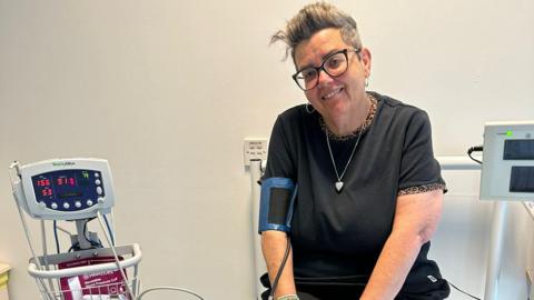 An older woman with short grey hair sits in a hospital room, attached to a heart rate monitor machine. She wears glasses, silver hooped earrings, a black t-shirt with a leopard-print trim around the sleeves and neckline.