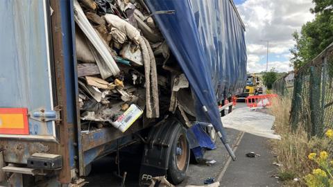 View of the side of the large HGV trailer, covered in blue plastic, with a triangular-shaped opening to the side where rubbish is piled high and falling out on to the pavement