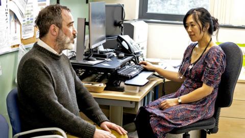 A man in a grey jumper talks to a female GP in a red and blue dress in a room in a doctor's office