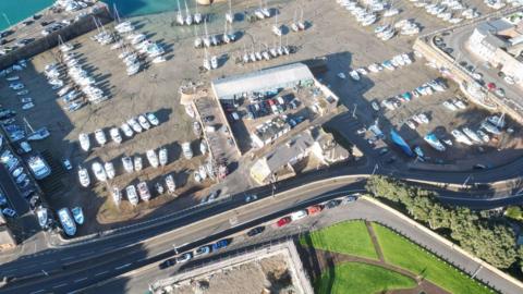 A drone photo of the La Folie Inn site in St Helier Harbour. The tide is out and there are many boats laid up, including yachts, dinghies and motorboats. There are cars parked by one of the buildings and there is a road and grassy park opposite the site.