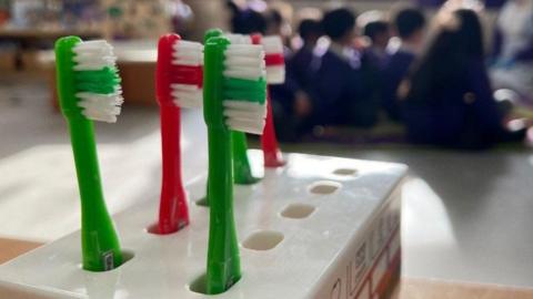 In the foreground there is a toothbrush holder. Three green and two red toothbrushes are visible. Children, who are blurred, are sitting in the background.