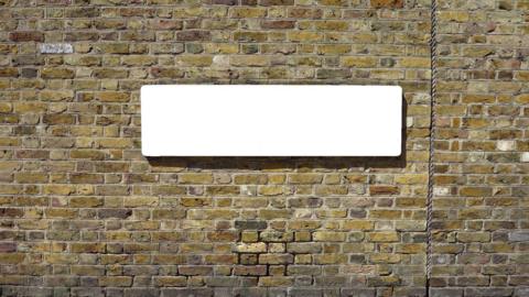 A blank road sign on a brick wall. The bricks are yellow and brown in colour. The road sign is a blank white slate where a name would usually be. 