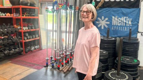 An older woman wearing a pink T-Shirt with glasses standing in front of gym equipment 