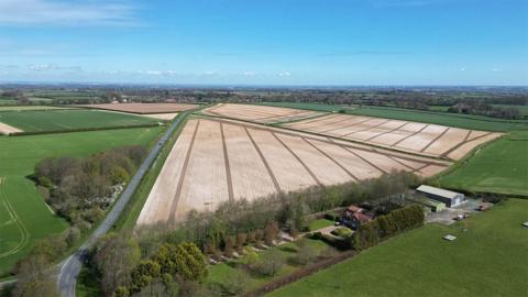 Aerial view of the proposed site near Beverley. It shows ploughed fields surrounded by hedges with a road running along the left. A farm with barns is at the bottom of the picture.