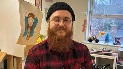 Aaron pictured with a black beanie hat on and has a long ginger beard. He is wearing a black and red plaid shirt and thin gold rimmed circular framed glasses. 