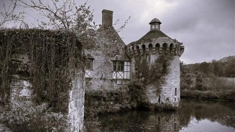 A stone castle from the Norman era of England. There is greenery and a moat beside the castle.
