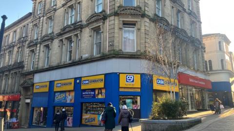 Blue and yellow signs adorn the front of multi-story building. Above the ground floor it is made of stone and had decorative features around the tall windows on each floor.