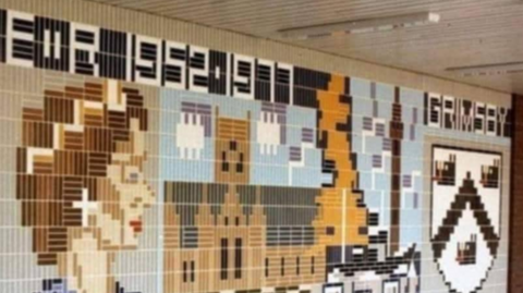 Photo of 1970's mosaic wall in Grimsby. It is multi-coloured and features images of a woman, a building and a map of the UK, as well as the town's Dock Tower and the town's crest.