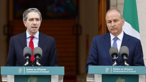 Taoiseach Simon Harris, to the left of the image, and Tánaiste Micheál Martin, to the right of the image. They are both standing behind green podiums with "Government of Ireland" written on the front in Irish and English. There are grey microphones on top of both podiums. Both men are wearing navy suits. Harris is also wearing a lilac shirt and a red tie. Martin is wearing a white shirt and a blue tie. 