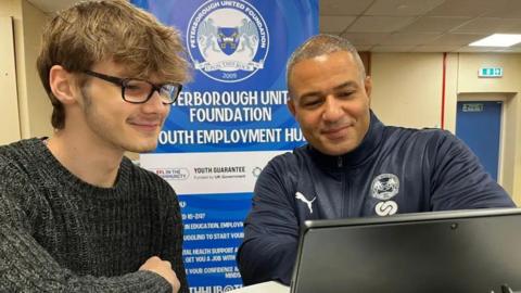 Young man wearing a grey jumper and glasses looking at a laptop with an older man with information about the employment hub behind them.
