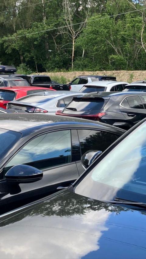 Lots of cars parked closely together with a fence and trees in the background