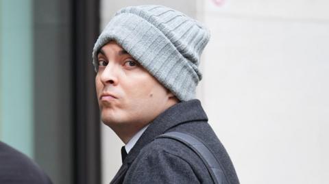 Che Homersham wearing a grey beanie and dark coat, glancing back while arriving at court.