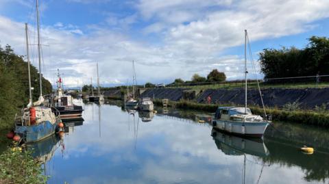 A series of boats are moored each side of the channel leading to Lydney Harbour. They are a mixture of small and medium-sized sailing boats and the water is clear and still.