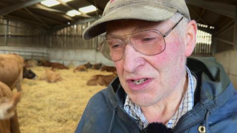 Eric Murley wearing a browncap and a blue jacket and checked shirt in a shed with livestock.