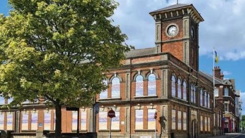 The exterior of the red brick town hall with its many windows bordered up and a flagpole outside the main entrance, with a large tree with green foliage in foreground