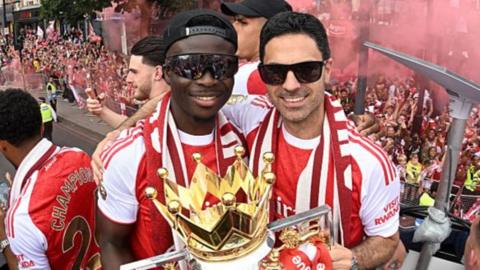 Bukayo Saka and Mikel Arteta, both in sunglasses and red and white team kit, with the Premier League trophy on Arsenal's bus parade