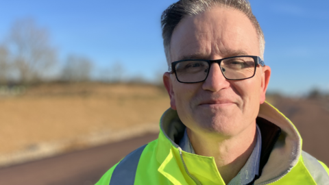 Chris Griffin is standing outside by a section of the A47 where work is under way. He is wearing a yellow fluorescent hi-vis jacket. He is also wearing glasses and is looking directly at the camera and smiling.