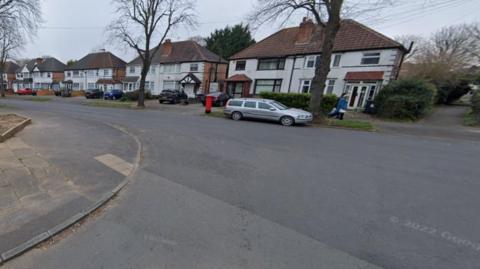 A generic image showing part of Scribers Lane going from left to right. A different road, at a junction with Scribers Lane, is in the foreground. One vehicle is parked opposite.