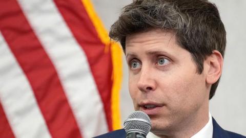 Sam Altman is speaking into a microphone, with the red stripes of the US flag visible in the background behind him.