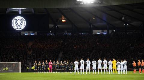 Two rows of football players applauding in front of a dark stadium filled with fans.