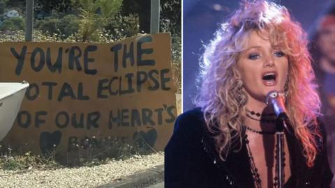 On the left A cardboard sign reads 'You're the total eclipse of our hearts Bonnie' On the right Bonnie Tyler singing into a microphone. She has big blonde hair and a black dress on. Behind is a drummer - he is slightly blurred, as Bonnie is the focus of the image.