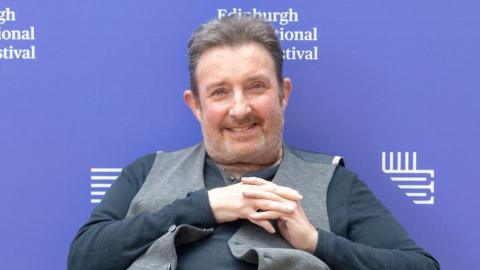 Denzil Meyrick smiles at the camera. He wears a dark grey top with a lighter grey waistcoat over the top. His hands are clasped across his chest and he is sitting down. A purple sign behind him reads Edinburgh International Festival.