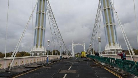 A view from the middle of the road across Albert Bridge. The clouds are thick  and quite dark. There are buildings and trees and and a highway maintenance van in the background.
