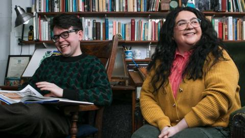 Inside the Alasdair Gray's archive, comedians Ashley Storrie and Christopher Macarthur-Boyd sit in front of a wall of bookshelves - Christopher, in a green and black patterned jumper, flips through an art book while Ashley, in green trousers, an orange shirt and a yellow cardigan, smiles off to the left.