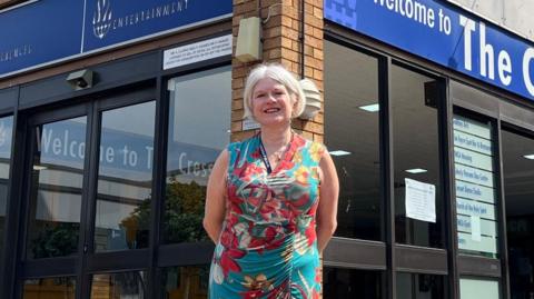 Louise Chantal is standing outside the Cresset building. It has brown bricks and electric glass doors at the entrance. She is smiling with her hands behind her back. 