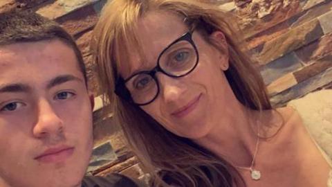 In this close up selfie image we see teenager Will Radford on the left and his mum Jane on the right. Will has short dark brown hair. Jane has long light brown hair with a fringe and wears glasses with black frames. She wears a necklace with a white pendant. 