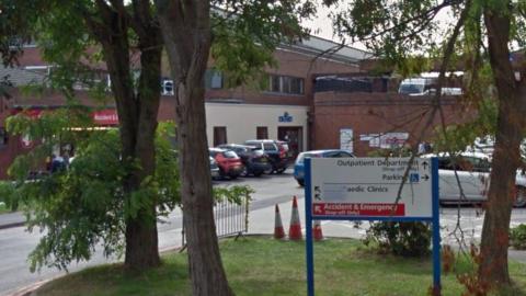 A Google street view of a circular grass verge with trees on it and a blue and white sign with a red 'accident and emergency arrow'. Behind it is a car park and a red entrance where you can see the word 'accident'.