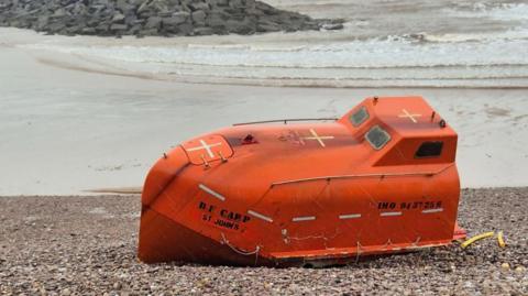 A small orange lifeboat on a pebble beach in Devon. It is orange, has writing on the front and back. There are three yellow crosses across the top of it, and two small windows at the top. The sea is behind it and is cloudy.