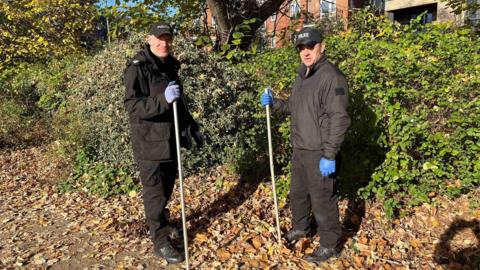 Two police officers with rubber clothes stand near a bush with sticks