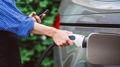 Woman with blue stripy shirt charging an electric car while looking at a mobile phone.