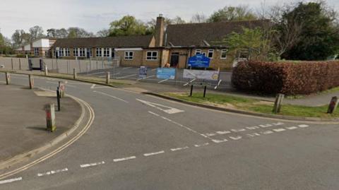 A single story pale brick building with a playground and parking spaces to the foreground, surrounded by wire fencing. A blue sign stands inside the school grounds, situated on a corner of a road.
