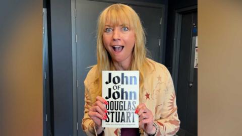 Sara Cox with her mouth open in surprise while holding up a book with the title, John of John by Douglas Stuart