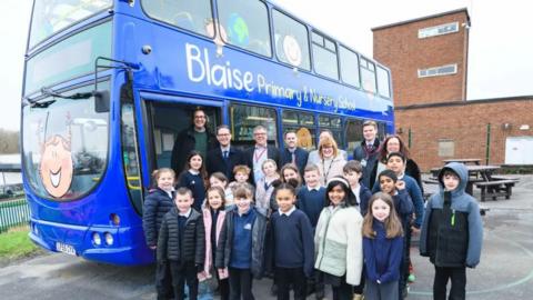 A group of children standing in front of a blue double decker bus, with the words 'Blaise Primary and Nursery School' written on its side