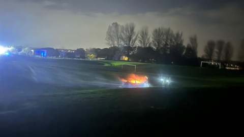 The picture, taken at dusk, shows a car on fire on the embarkment of a football pitch with two goalposts behind it. 