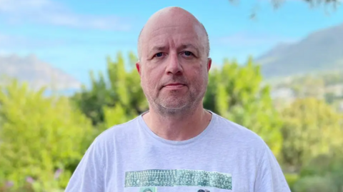 Man with a shaved head and light stubble stands outdoors in front of blurred greenery and distant mountains, looking directly at the camera with a neutral expression. He is wearing a light grey T-shirt, and the scene is brightly lit with natural daylight.