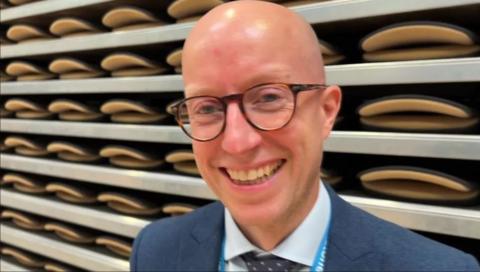 A man smiling while looking straight at the camera. He is wearing a navy suit with a blue shirt and tie and tortoiseshell glasses