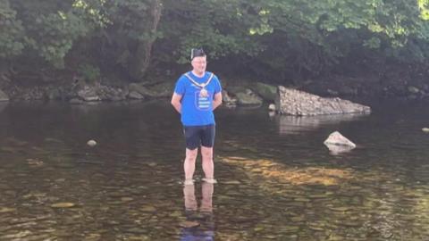 A man wearing a blue T-shirt, black shorts and mayoral chain and hat stands in a river.