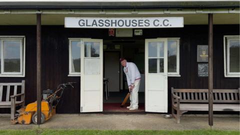 A man stands in the entrance of a club house which has a sign above which says 'Glasshouses CC'. He is wearing a cap and is dressed in cricket whites and holds a bat. There are benches of either side of the entrance and on the left is an orange lawn mower.  